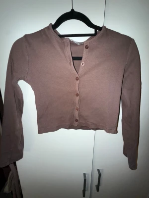 Brun cropped cardigan med knappar - Snygg brun cropped cardigan med långa ärmar och knäppning framtill. Tillverkad i ribbad bomullsmix som ger en skön och stretchig känsla. Perfekt att styla över ett linne eller t-shirt för en avslappnad look.