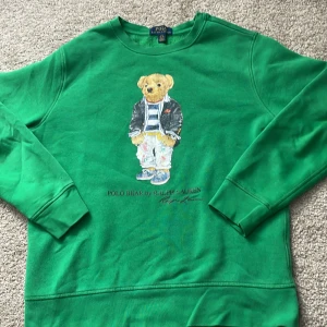 Grön sweatshirt Polo Bear Ralph Lauren - Grön sweatshirt från Ralph Lauren med det ikoniska Polo Bear-motivet på bröstet. Tröjan har rund halsringning, långa ärmar och ribbade muddar. Perfekt för dig som gillar streetwear och vill ha en klassisk men lekfull look.