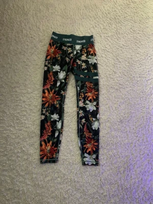 Blommiga leggings från Stronger - Snygga leggings från Stronger med färgglatt blommönster i orange, vitt och grönt på mörk botten. Tillverkade i återvunnen polyester och elastan för en stretchig och bekväm passform. Perfekta för dig som vill sticka ut på gymmet eller i vardagen.