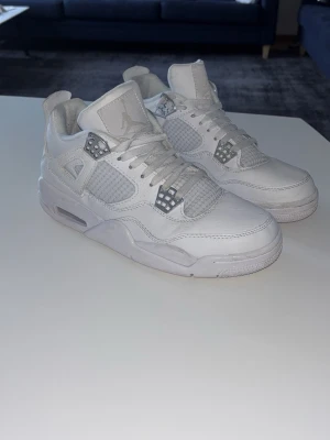 Nike Air Jordan 4 Pure Money - Nike Air Jordan 4 sneakers i helvitt skinn med silverdetaljer och klassisk Jumpman-logga på hälen. Skorna har snörning, meshpaneler på sidorna och en chunky sula för extra streetstyle-vibe. Perfekta för dig som gillar ikoniska sneakers med clean look.