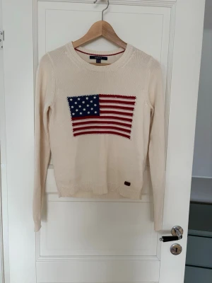Beige stickad tröja med USA-flagga GANT - Stickad beige tröja från GANT med rund halsringning och lång ärm. På bröstet finns en stor broderad amerikansk flagga i blått, rött och vitt. Tröjan har ribbade muddar och en klassisk passform.