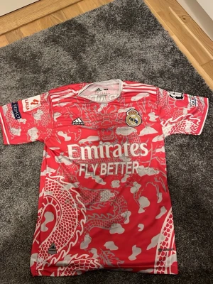 Real Madrid rosa matchtröja Adidas - Snygg rosa Real Madrid matchtröja från Adidas med drakmönster i vitt och grått. Tröjan har klubbmärke, Adidas-logga och sponsortryck på bröstet samt tryck på ärmarna. Tillverkad i lätt och ventilerande material.