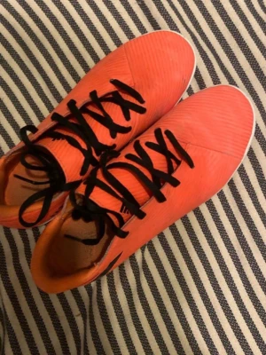 Adidas Nemesis orange fotbollsskor - Säljer ett par Adidas Nemesis fotbollsskor i en snygg orange färg med svarta snören och vit sula. Skorna har en sportig design och är perfekta för dig som vill sticka ut på planen. Tillverkade i syntetmaterial för bra komfort och grepp.