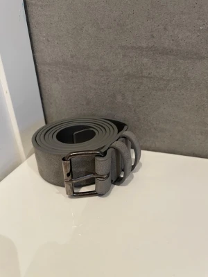 Grått bälte i mockaimitation - Snyggt grått bälte i mockaimitation med stilren metallspänne. Bältet har en enkel och modern design som passar till många olika outfits. Perfekt accessoar för att lyfta din look.