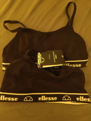 Svarta bralettes från Ellesse - Två svarta bralettes från Ellesse med smala axelband och elastiskt band nedtill med vit logga och text. Snygg och sportig design, perfekta under tröjor eller till vardags. Mjukt och stretchigt material för skön passform.