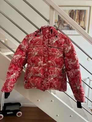 Röd Peak Performance dunjacka - Snygg röd dunjacka från Peak Performance med vitt blommigt mönster. Jackan har dragkedja, snörning i huvan och långa ärmar med svarta muddar. Perfekt för kalla dagar och vintersäsongen.
