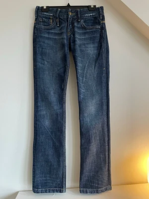  Blå lågmidjade jeans straight fit -  blå HVANA jeans med rak passform, låg midja och fickor. Jeansen har en snygg tvättad look och är tillverkade i slitstarkt bomullsdenim. Perfekta för dig som gillar tidlös stil och vill ha ett par jeans som funkar till allt. Mått: midja- 37,5 cm     Innerbenslängd - 85 cm