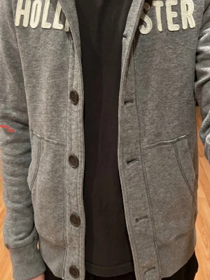 Grå hoodie från Hollister😁😎 - Snygg grå hoodie från Hollister med knappar och huva. Mjuk insida i fleece och ribbade muddar. Perfekt för en avslappnad och stilren look. Klassisk Hollister-logga på insidan. Sällsynt hollister hoodie. Storlek är M och priset går att tala om😁😁