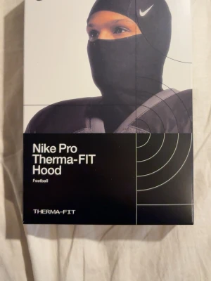 Nike Pro Therma-FIT Hood svart - Nike Pro Therma-FIT Hood i svart med silverfärgad Nike-logga. Balaklava i sportig design som täcker huvud, hals och nacke. Tillverkad i värmande syntetmaterial, perfekt för kalla dagar och träning utomhus.