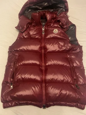 Vinröd dunväst från Moncler - Säljer en vinröd dunväst från Moncler med glansig finish och klassisk Moncler-logga på bröstet. Västen har huva, två dragkedjeförsedda fickor och quiltad design. Perfekt för lager-på-lager under kyliga dagar.