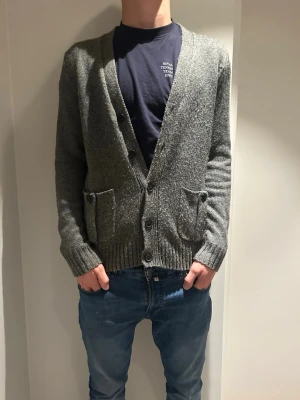 Grå stickad kofta med mockadetaljer - Säljer en grå stickad kofta med knappar framtill och två fickor. Koftan har bruna mockalappar på armbågarna och axelremmar för en cool touch. Perfekt för lager-på-lager under höst och vinter.Modellen är 187cm lång, hör av er vid frågor 😀