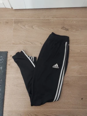 Svarta Adidas träningsbyxor med dragkedja - Säljer ett par svarta träningsbyxor från Adidas med vita ränder längs sidorna och logga på benet. Byxorna har resår och snörning i midjan samt dragkedja vid bensluten. Perfekta för träning❣️