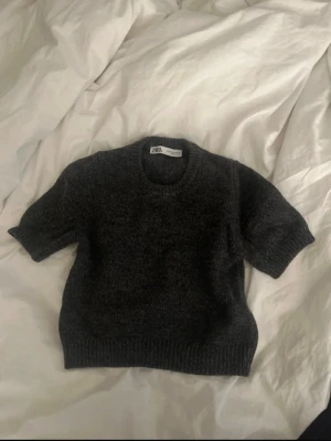 Mörkgrå stickad tröja från Zara - Säljer en mörkgrå stickad tröja från Zara i storlek XS. Tröjan har korta ärmar, rund halsringning och ribbade muddar vid ärmslut och nederkant. Perfekt för lager på lager under hösten eller våren. Aldrig använd