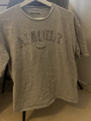 Grå t-shirt Zadig & Voltaire med paljetter - (Har tappat diamanter )Säljer en grå t-shirt från Zadig & Voltaire med coolt paljettmönster som stavar 'AMOUR' och en liten vingdetalj under texten. T-shirten har korta ärmar och är gjord i mjuk bomull. Perfekt för dig som gillar statement-plagg med lite extra bling. 