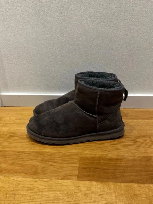 Mörkgrå UGG - Säljer ett par mörkgrå UGG boots med mjuk päls på insidan och platt sula. Skorna är tillverkade i mocka och har en rund tå. Perfekta för kalla dagar och har klassisk UGG-logga baktill.