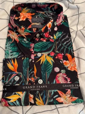 Blommig skjorta från Grand Frank - Säljer en oanvänd skjorta från Grand Frank. Skjortan har svart botten med stora blommor i orange, rosa, grön och gul. Klassisk krage och knappar framtill. Perfekt för dig som vill sticka ut med en unik look. Storlek är L och passform är slim fit.