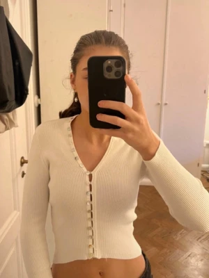 Vit ribbad kofta med pärlemorknappar - Säljer en croppad, vit ribbad kofta med v-ringning och snygga pärlemorknappar hela vägen framtill. Långärmad och figurnära passform