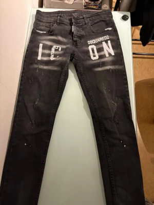 Svarta Dsquared2 ICON slim jeans - Säljer ett par svarta Dsquared2 ICON slim jeans med vit text framtill och slitna detaljer. Jeansen har klassisk femficksmodell, röda sömmar på insidan och är tillverkade i bomull. Snyggt streetwear-plagg med smal passform och coola tryck.