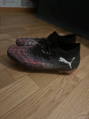 Puma fotbollsskor i svart och orange - Säljer ett par snygga Puma fotbollsskor med svart och orange mönster, vita Puma-loggor på sidorna och snörning. Ovandelen är i syntetmaterial och skon har en strumpliknande passform runt ankeln. Sulan är orange med fasta dobbar för gräsplan.