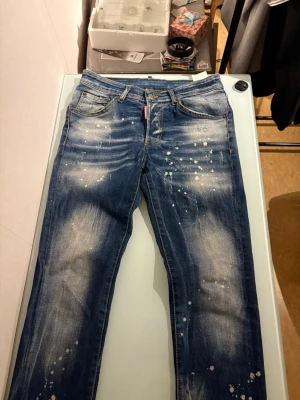 Dsquared2 blå slim jeans stl 42 - Säljer ett par blå slim fit jeans från Dsquared2 i storlek 42. Jeansen har coolt slitna detaljer och vita färgstänk för en edgy look. Klassisk femficksmodell med gul kontrastsöm och knappgylf. Tillverkade i Italien i robust denim.
