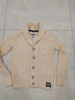 Beige kofta från Superdry XL - Säljer en beige kabelstickad kofta från Superdry i storlek XL. Koftan har stora knappar framtill, bred krage och ribbade muddar. Perfekt för lager-på-lager och har en klassisk look med snygg detalj vid nederkanten.