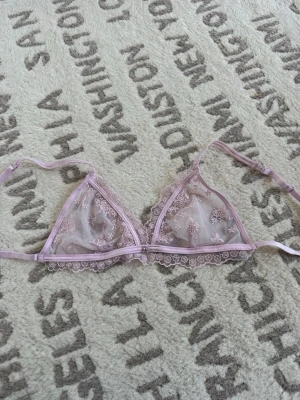 Ljusrosa spetsbralette med rosett - Söt ljusrosa bralette i mesh och spets med broderade blommor och små rosetter. Justerbara axelband och dekorativ liten metallrosett framtill. Transparent och feminin design med fina spetskanter.