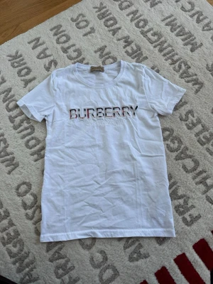 Vit Burberry t-shirt med logga - Snygg vit t-shirt från Burberry med klassisk logga broderad framtill i svart, rött och vitt. T-shirten har rund halsringning och korta ärmar. Tillverkad i mjuk bomull som känns skön mot huden. Perfekt för en clean och stilren look.