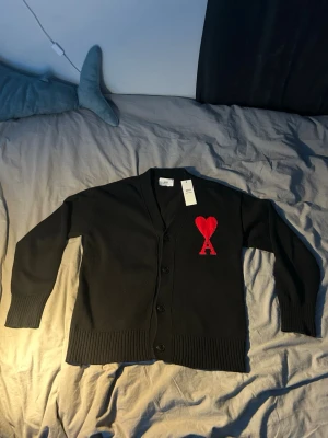Svart kofta från Ami med rött hjärta - Säljer en svart kofta från Ami Alexandre Mattiussi i storlek M. Koftan har ett stort rött hjärta med bokstaven A broderat på bröstet, ribbade muddar och knäppning framtill. Perfekt för dig som vill ha en stilren men ändå unik look.