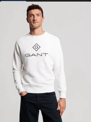 Vit sweatshirt från GANT - Säljer en stilren vit sweatshirt från GANT i bomull med svart broderad logga framtill. Tröjan har rund halsringning, långa ärmar och ribbade muddar. Perfekt för en clean och klassisk look.