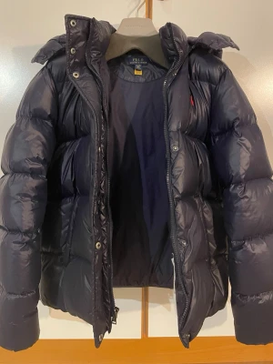 Mörkblå pufferjacka Polo Ralph Lauren - tjej - Säljer en mörkblå pufferjacka från Polo Ralph Lauren. Priset kan diskuteras. Jackan har huva, dragkedja och tryckknappar framtill samt två fickor. Klassisk röd logga på bröstet och riktigt skön vaddering som håller dig varm under kalla dagar. Storlek: 	•	Märkt XL / TG (16 år) 	•	Ungdomsstorlek (största barnstorleken) 	•	Motsvarar cirka XS vuxenstorlek