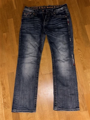 Rock Revival blå jeans med broderi - Säljer ett par Rock Revival jeans i blå tvätt med tydliga slitningar och kontrastsömmar. Jeansen har raka ben/lite bootcut, broderade bakfickor med dekorativa detaljer och metallknappar. Materialet är klassisk jeansbomull och passformen är normal. Snygga detaljer och cool look! Storlek är 32 längd 32 midja. Extremt liten fläck på ett av låren som ej syns. Skriv för mer bilder så kan vi diskutera pris.