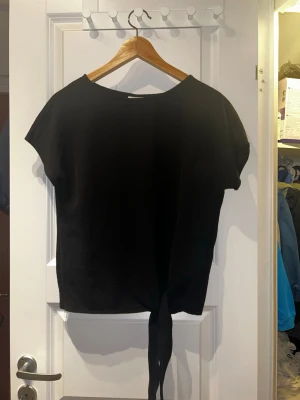 Svart knytblus från Vila - En svart blus från Vila med korta ärmar och rund halsringning. Toppen har en snygg knytning nedtill som ger en cool twist till looken. Tillverkad i ett mjukt och lätt material som känns skönt mot huden.