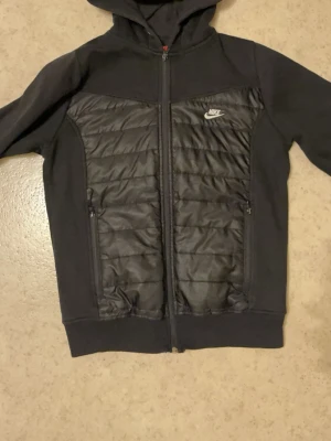 Svart pufferjacka från Nike med luva  - Säljer en svart pufferjacka från Nike med huva och hel dragkedja. Jackan har quiltade partier fram och bak, samt två fickor med dragkedja. Nike-logga på bröstet. Perfekt för kyliga dagar och riktigt snygg streetstil. Storlek xs 