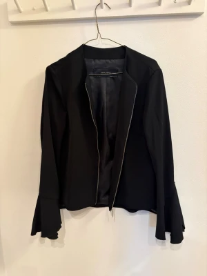 Svart kavaj med volangärm från Zara - Säljer en svart kavaj från Zara Basic med snygga volangärmar och dragkedja framtill. Jackan har en stilren och enkel design med lång ärm och lätt figursydd passform. Perfekt för dig som vill ha en trendig och clean look.