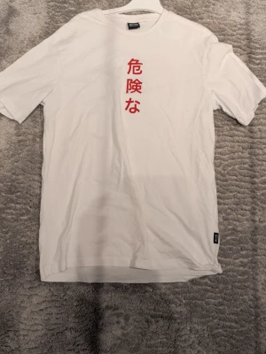 Vit t-shirt med draktryck och japansk text - Säljer en vit t-shirt från Only & Sons med röd japansk text framtill och en stor svart drake med röd sol på ryggen. T-shirten har korta ärmar och är gjord i mjuk bomull. Perfekt för dig som gillar streetwear och asiatiska influenser.