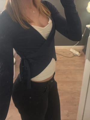 Mörkblå omlottopp med knyt - Snygg mörkblå omlottopp med långa ärmar och knyt i sidan. Toppen har en croppad passform och djup v-ringning, perfekt att styla med ett linne under. Materialet känns mjukt och följsamt. Gina young 170