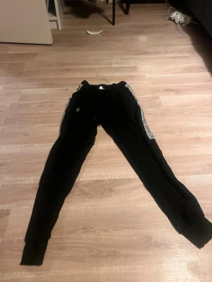 Svarta mjukisbyxor Maya Delorez 2XS - Svarta mjukisbyxor från Maya Delorez i storlek 2XS. Byxorna har gråa sidor med texten 'Maya Delorez' längs benen och snörning i midjan. Tillverkade i mjukt material, perfekta för chill dagar eller träning.