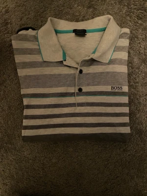 Randig grå BOSS piké  - Snygg randig piké från Hugo Boss i olika grå toner med inslag av svart och turkos. Klassisk krage med två knappar och BOSS-logga broderad på bröstet. Perfekt för dig som gillar stilrena och moderna plagg.