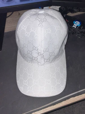 Vit Gucci keps - Säljer en stilren vit keps från Gucci med det klassiska GG-mönstret över hela kepsen. Kepsen är tillverkad i bomull och har justerbart spänne bak i vitt läder. Perfekt accessoar för att lyfta din outfit. Hör av er i dm tyckte den inte satt gött på mig så säljer den då jag fick den av min bror.