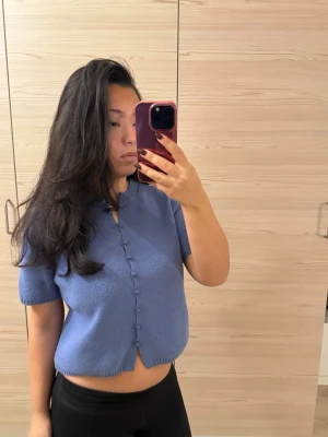 Blå kortärmad kofta med knappar - Säljer en söt blå kofta med korta ärmar och rund hals. Koftan har dekorativa knappar framtill och är stickad i ett mjukt material. Modellen är croppad och har ribbade kanter vid ärmslut och nederkant. Perfekt för vår och sommar!