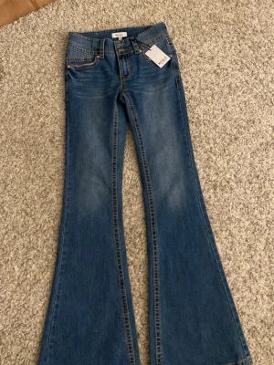 Blå bootcut jeans från RYVLS - Säljer ett par snygga blå bootcut jeans från RYVLS. Jeansen har klassisk femficksdesign, markerade sömmar och två bakfickor med knappdetalj. Tillverkade i jeansmaterial med en mellanblå tvätt, helt ny i storlek 164!💗