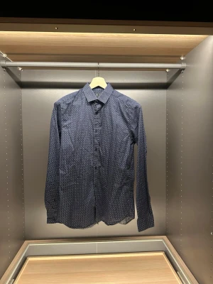 Mörkblå slim fit skjorta från Riley - Säljer en mörkblå slim fit skjorta från Riley med små vita och ljusblå prickar. Skjortan har klassisk krage, långa ärmar och knäppning framtill. Perfekt för dig som gillar stilrena detaljer och smal passform.