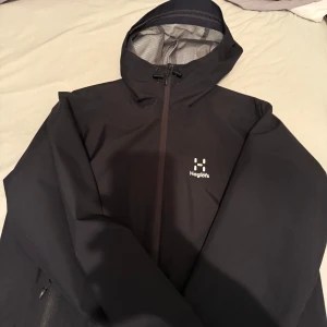 Svart vindjacka från Haglöfs - Säljer en svart vindjacka från Haglöfs med huva och dragkedja. Jackan har en stilren design med diskret logga på bröstet. Perfekt för blåsiga dagar och utomhusaktiviteter. Inuti finns ett nätfoder för extra komfort. Jackan är i Storlek M i dam men passar Man XS 800kr snabb affär 