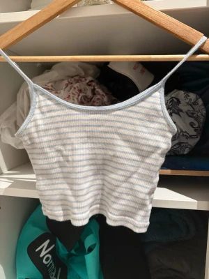 Ljusblå- och vitrandig linnetopp - Säljer en söt ribbad linnetopp med tunna axelband i ljusblått och vitt. Toppen har ett smalt randigt mönster och är croppad i modellen. Perfekt för varma dagar och enkel att matcha med jeans eller kjol.