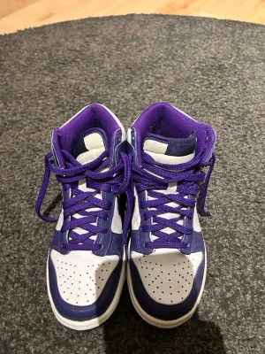 Nike Dunk High vit/lila sneakers - Nike Dunk High sneakers i vitt och lila med klassisk Swoosh på sidan. Skorna har snörning, perforerad tå och hög siluett. Tillverkade i skinn med matchande lila innerfoder och sula. Perfekt för dig som vill ha en clean och sportig look.