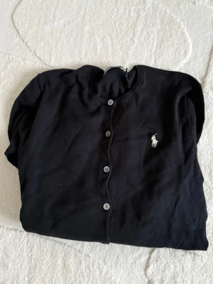 Svart kofta från Ralph Lauren - Klassisk svart kofta från Ralph Lauren med rund halsringning och knappar framtill. Tröjan har lång ärm och den ikoniska vita loggan broderad på bröstet. Perfekt att slänga över en t-shirt eller linne för en clean look.