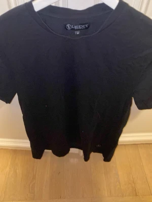 Svart t-shirt från LAGENCY - En klassisk svart t-shirt från LAGENCY i storlek M. T-shirten har rund halsringning och korta ärmar. Tillverkad i mjuk bomull som känns skön mot huden. Perfekt basplagg att ha i garderoben.