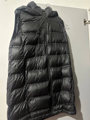 Svart lång dunväst med huva - Säljer en svart lång dunväst med huva och dragsko. Västen har quiltad design, dragkedja framtill och är ärmlös. Perfekt för lager på lager och riktigt snygg till höst- och vinteroutfits.