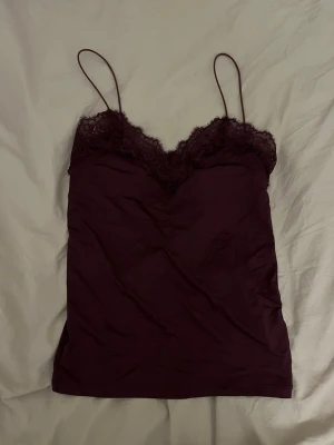 Vinröd spetsdetaljerad linne - Säljer en vinröd linnetopp med tunna axelband och vacker spets längs urringningen. Toppen har en djup v-ringning och är tillverkad i ett mjukt tyg som känns skönt mot huden. Perfekt att styla med jeans eller kjol för en trendig look.