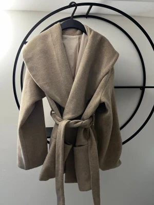 Beige omlottkappa med bälte - Säljer en beige kappa med bred krage och omlottmodell. Jackan har ett matchande bälte i midjan och stora fickor framtill. Materialet känns mjukt och lite ulligt, perfekt för kyligare dagar. Snygg och enkel att styla till olika outfits.